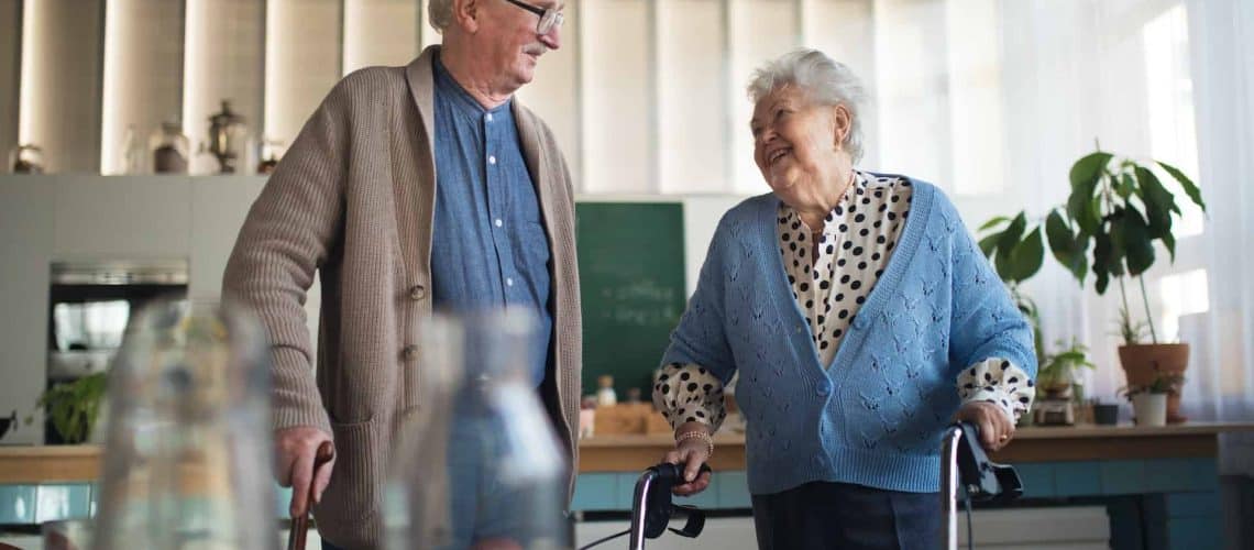 Elderly man and woman using walkers in a kitchen, smiling at each other, with plants and sunlight in the background.