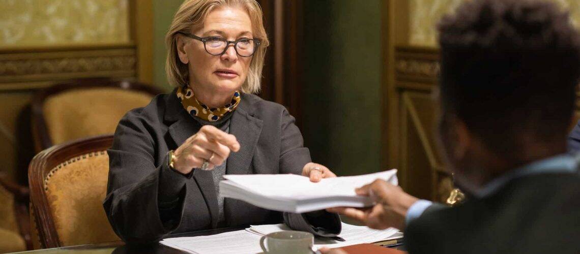 A woman with glasses hands a stack of documents to a man across a table in a formal setting.
