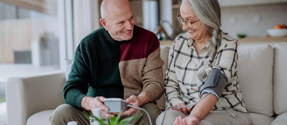 Elderly man helps woman measure her blood pressure with a monitor while sitting on a sofa indoors.