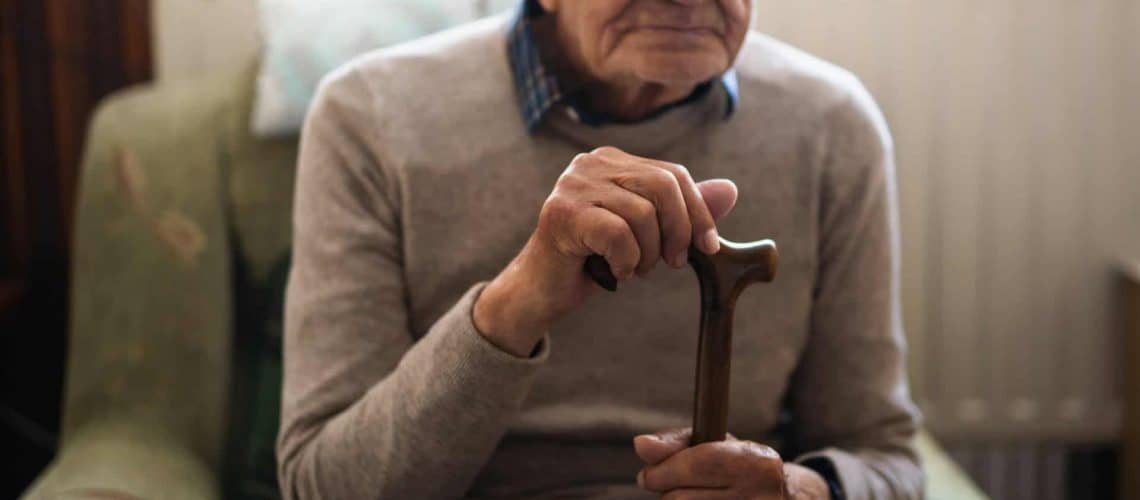 Elderly person sitting in an armchair, holding a wooden cane with both hands. The individual is wearing a gray sweater over a checked shirt.