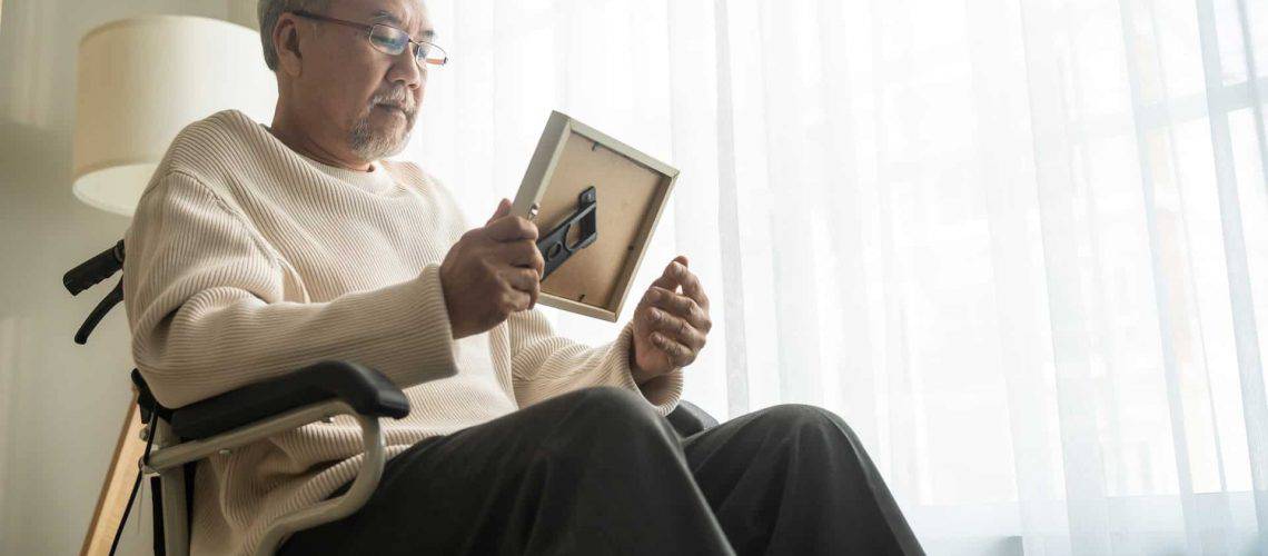 An elderly person in a wheelchair looks at a framed photo by a sunlit window.