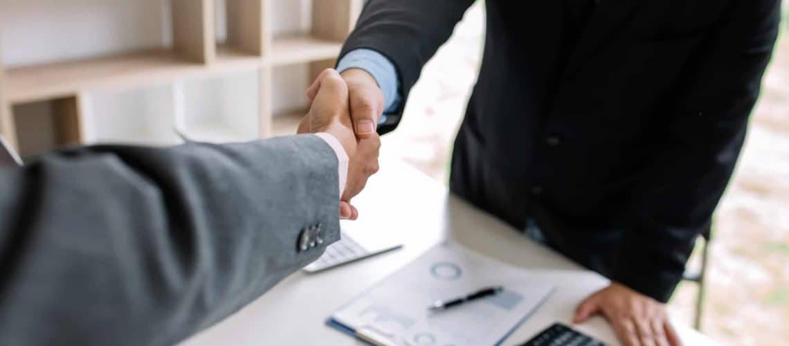 Two people in suits are shaking hands over a table with documents, a laptop, and a calculator.