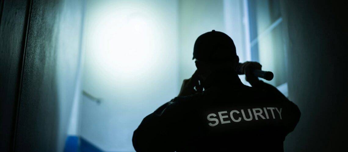 Security guard in a dimly lit hallway holds a flashlight and talks on a phone, with "SECURITY" written on the back of their jacket.