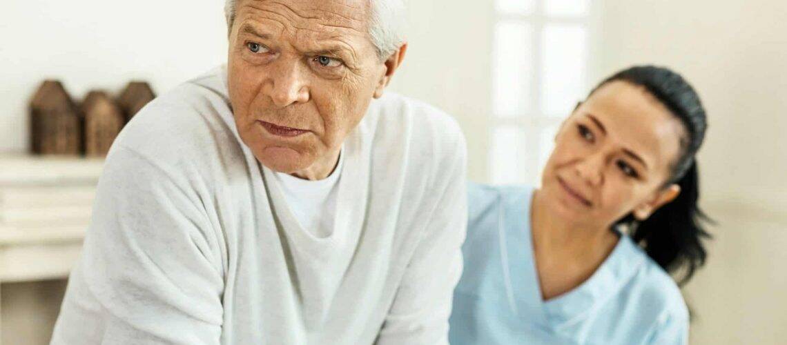 An older man in a white sweater looks away, sitting beside a woman in a blue uniform who appears to be a caregiver, in a bright room.