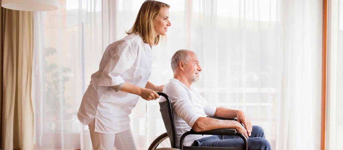 A woman stands behind a man in a wheelchair, both looking out a sunlit window with sheer curtains.