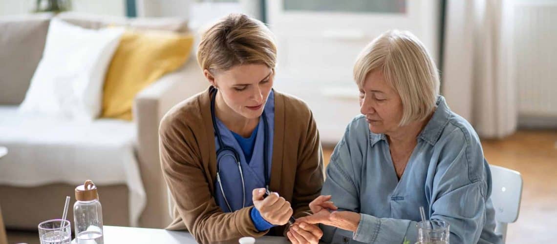 A healthcare professional discusses medication with an elderly woman at a table in a well-lit living room.