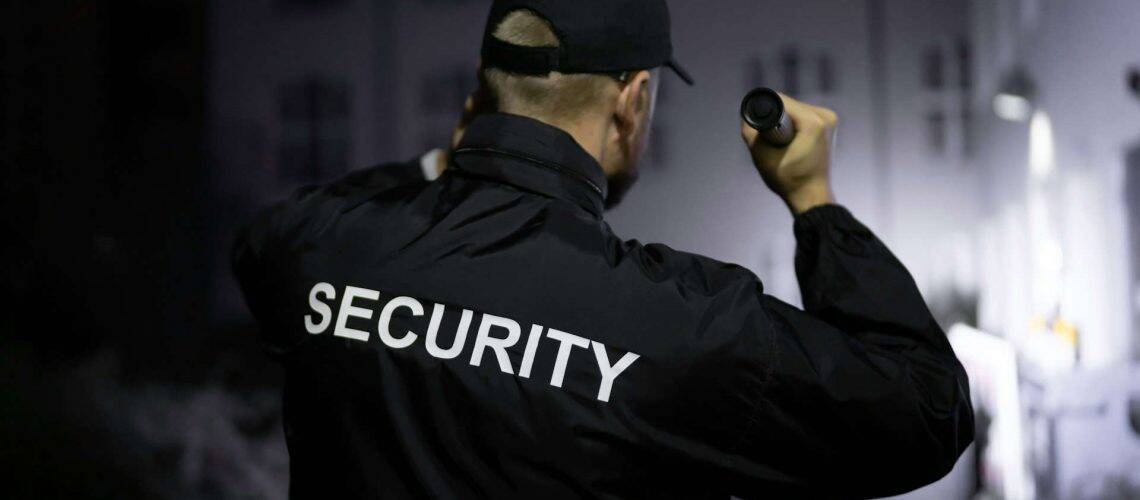 A person in a jacket with "SECURITY" on the back holds a flashlight, standing in a dimly lit area.