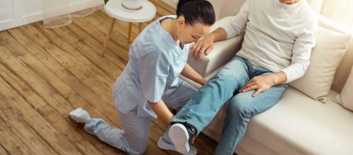 A healthcare professional kneels and examines the leg of a person sitting on a couch in a well-lit room.