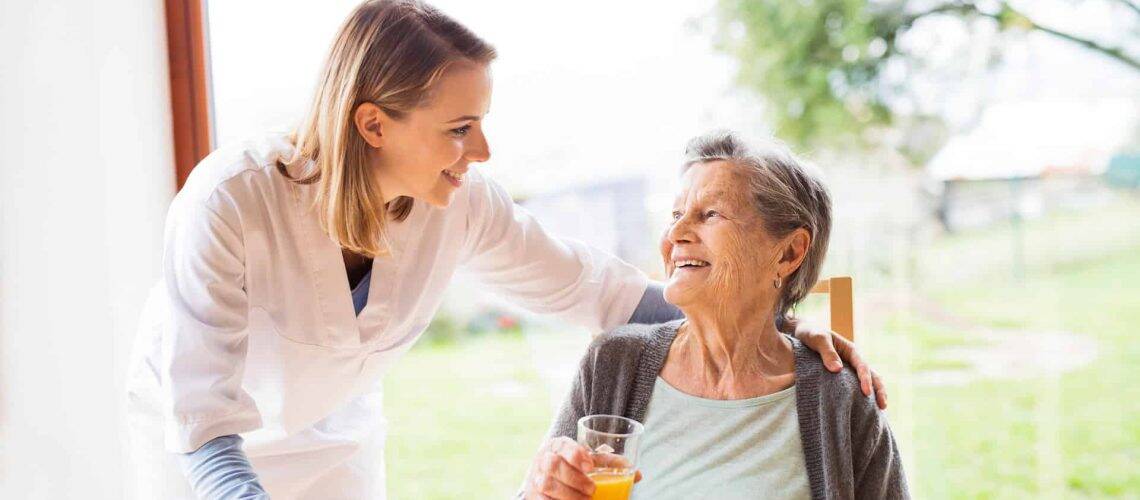 A caregiver smiles at an elderly woman holding a glass of orange juice. They are in a bright room with a view of greenery outside.