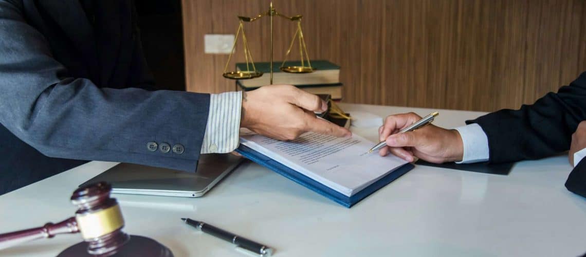 Two people in suits sitting at a desk; one points at a document while the other signs it. A gavel, pen, books, and scales of justice are visible in the background.