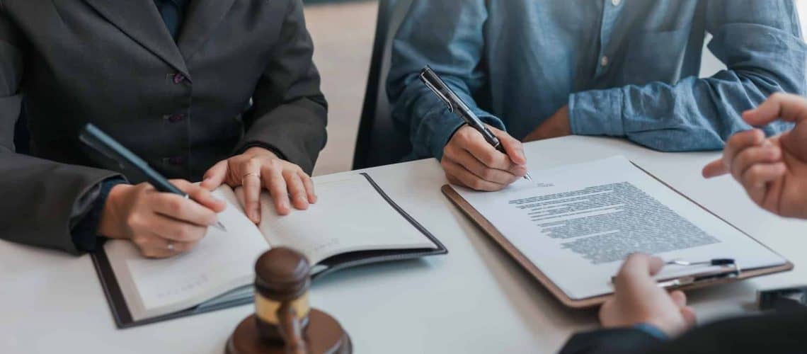 Two people signing documents at a table with a gavel, book, and a pen in hand, suggesting a legal or formal setting.