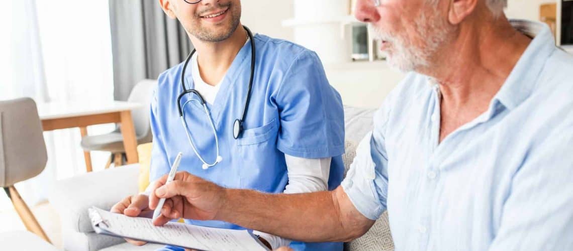 A healthcare professional in scrubs talks to an older man, who points to a document on a clipboard.