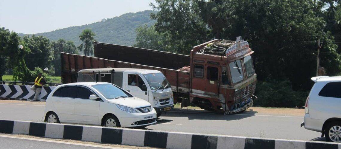 Trucks and cars are on a road by a green landscape. One truck appears to have fallen partially off the road.