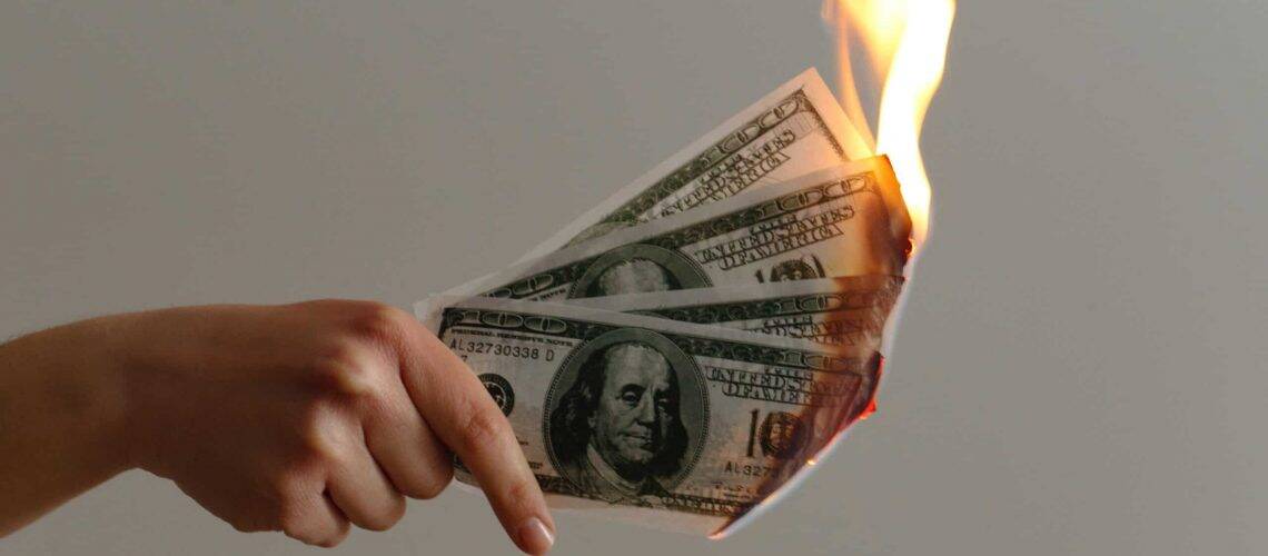 Hand holding burning hundred-dollar bills with flames on the right side against a plain background.