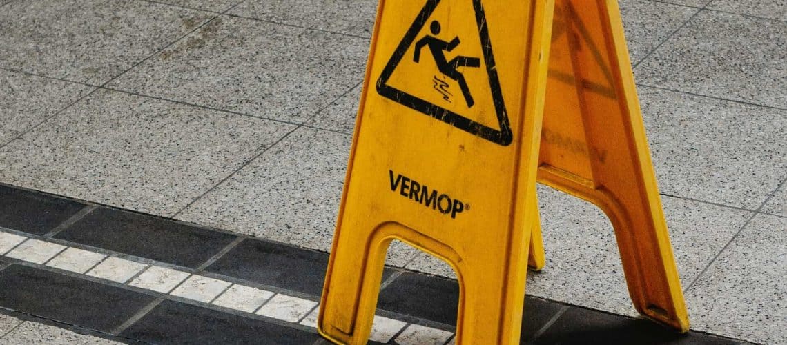 Yellow caution sign on a tiled floor warning of a wet surface with a slipping figure icon.