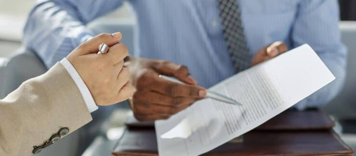 Two people in business attire reviewing a document, holding pens. One points at the paper, while the other listens attentively.