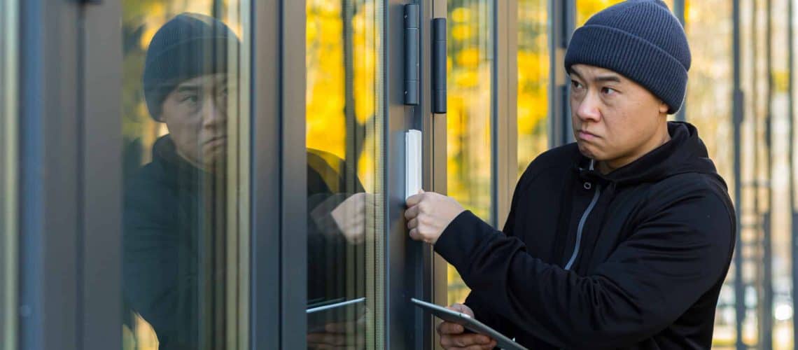Person wearing a beanie and black jacket attempting to open a glass door with a device. Reflections are visible in the glass.