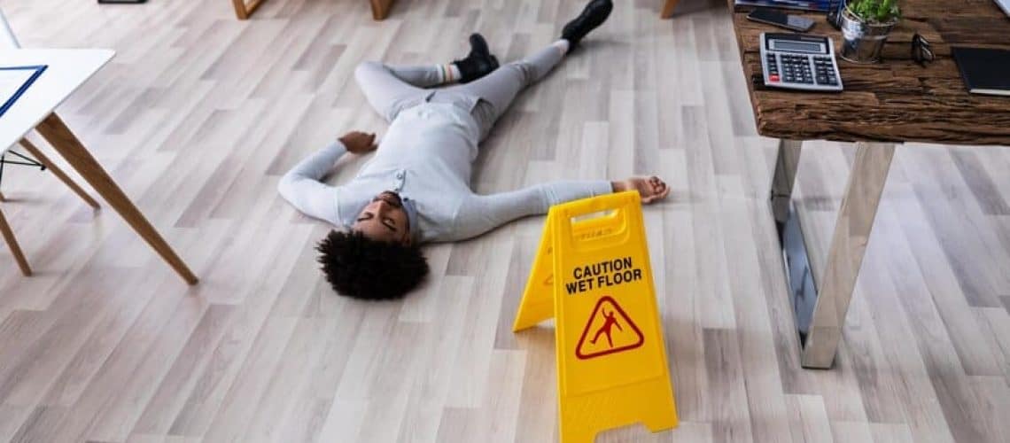 Person lying on the floor next to a "Caution Wet Floor" sign in an office setting.