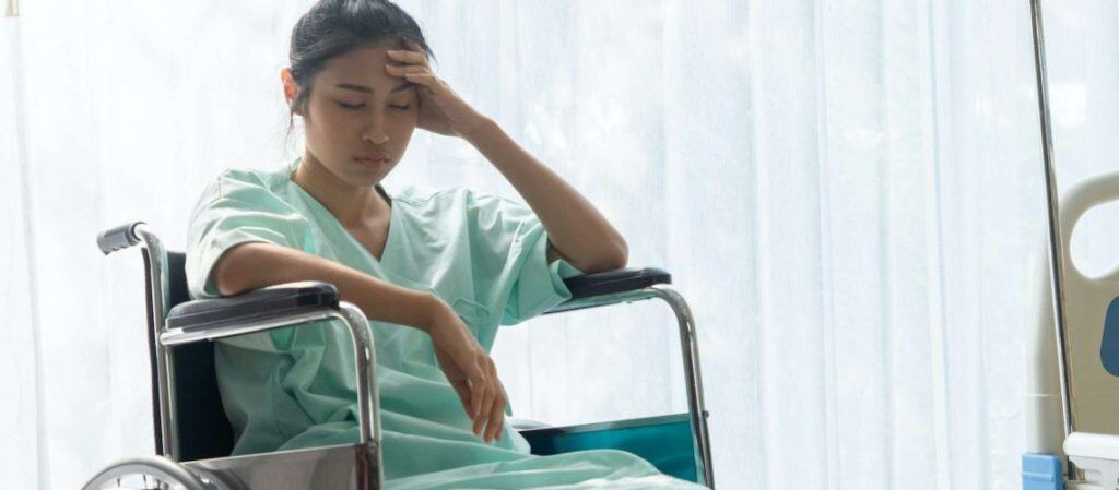 A woman in a hospital gown sits in a wheelchair by a window, resting her head on her hand and appearing contemplative or tired.