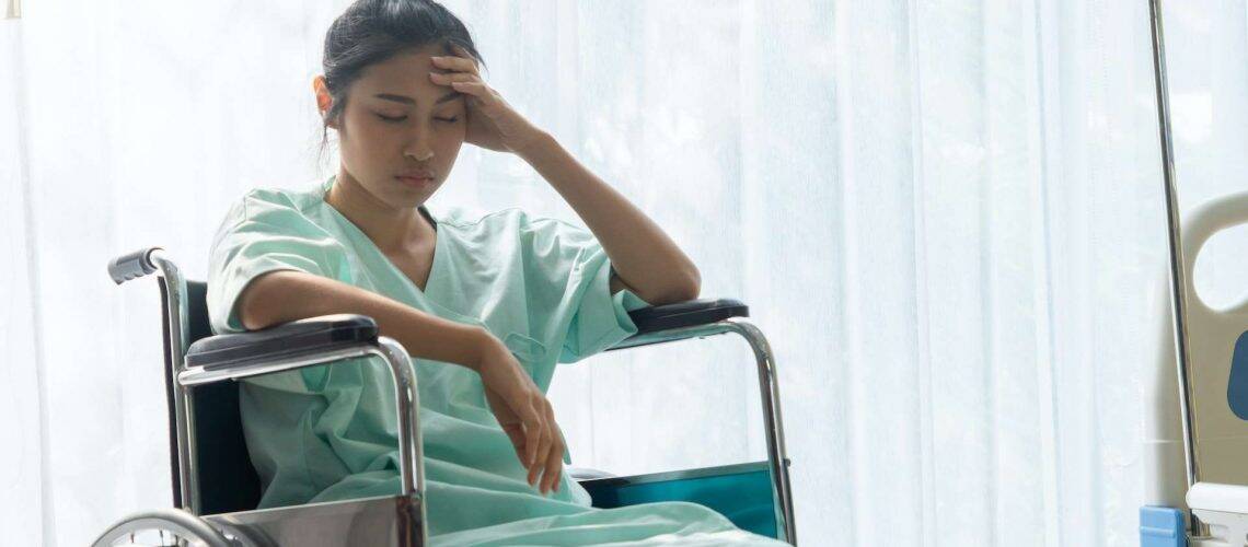A woman in a hospital gown sits in a wheelchair by a window, resting her head on her hand and appearing contemplative or tired.