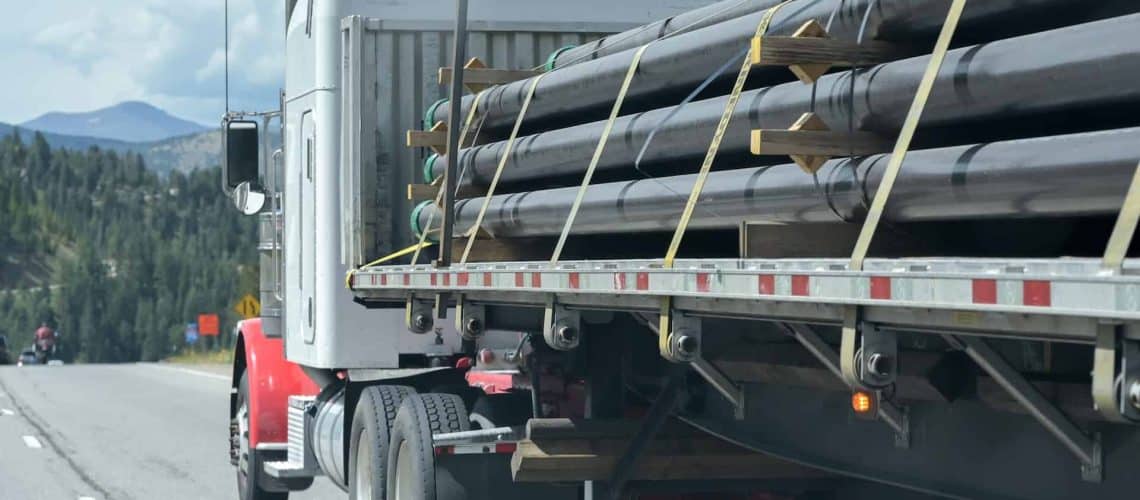 A truck hauls large pipes on a flatbed trailer along a highway, set against a backdrop of trees and mountains.