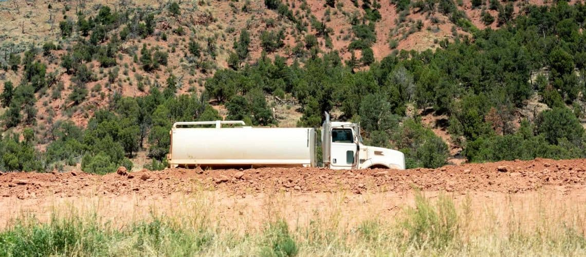 A white tanker truck is parked on a dirt road in a landscape with red rocky hills and scattered green trees.