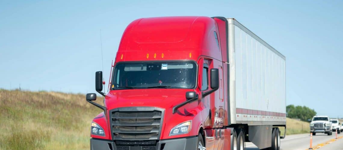 A red semi-truck travels on a highway with a gray trailer, clear blue sky in the background, and some grass on the roadside.