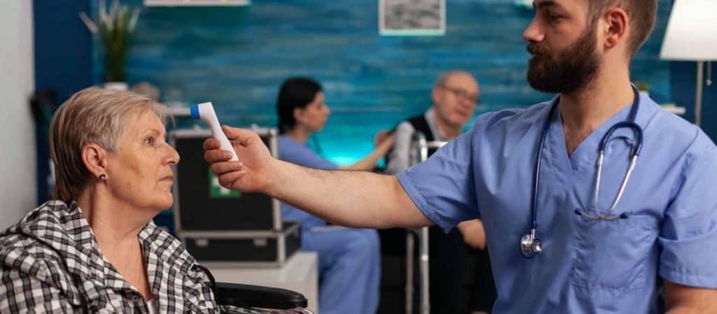 A healthcare worker in blue scrubs takes the temperature of an elderly woman in a clinical setting, while other medical staff and patients are visible in the background.