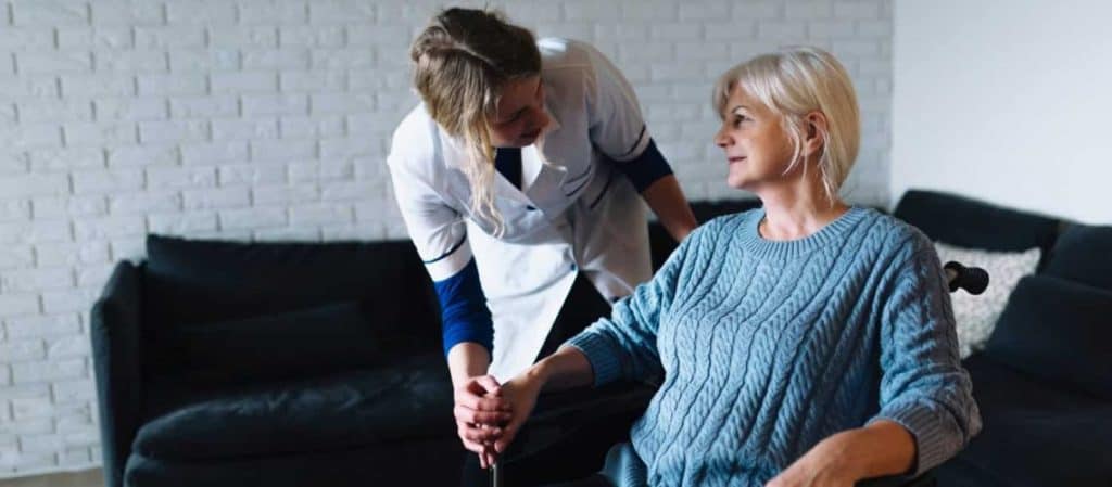 A caregiver in a white uniform leans over to assist an older woman sitting in a wheelchair in a living room.