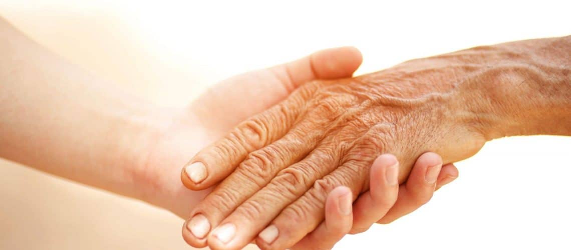 A younger person's hand supports an older person's hand against a plain background.