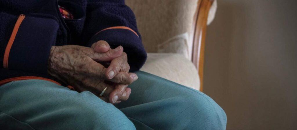 Elderly person with folded hands sitting on a chair, wearing a blue jacket and green pants.