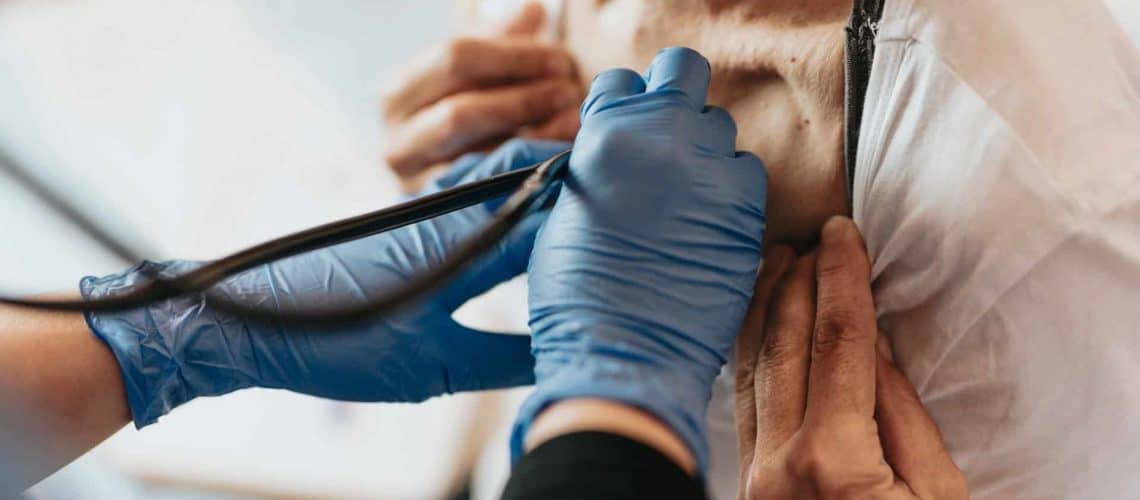 Person wearing blue gloves checks a patient's pulse using a stethoscope on their back.