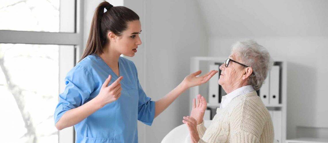 A nurse in blue scrubs gestures while talking to an elderly woman in a white sweater and glasses in a brightly lit room.