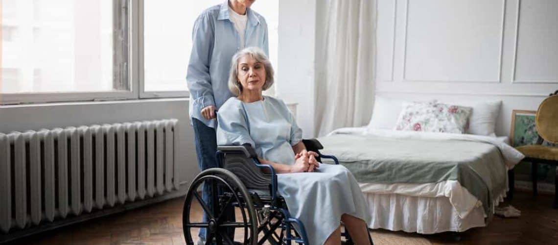 An elderly woman sits in a wheelchair near a bed, with a person standing behind her in a well-lit room.