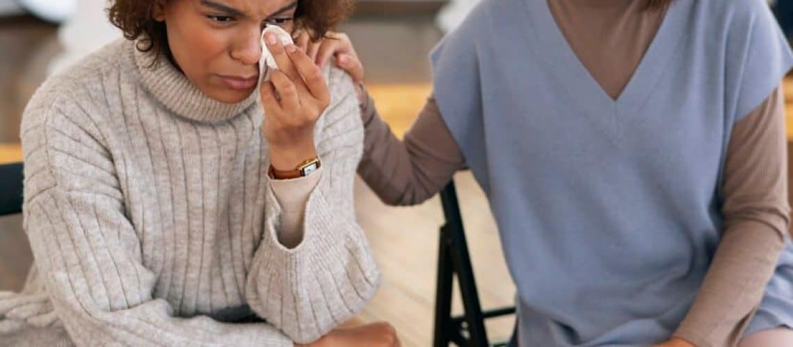 A person in a sweater is sitting and wiping tears with a tissue, while another person beside them, in a blue vest, offers comfort with a hand on their shoulder.