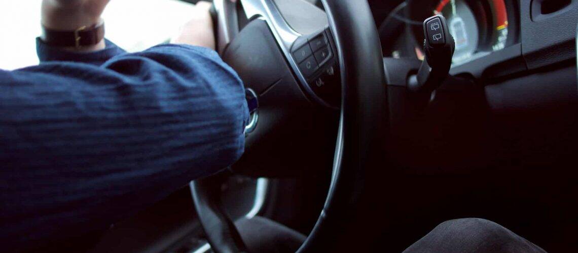 Close-up of a person wearing a blue shirt, steering a car. The focus is on the steering wheel, dashboard, and gearshift lever.
