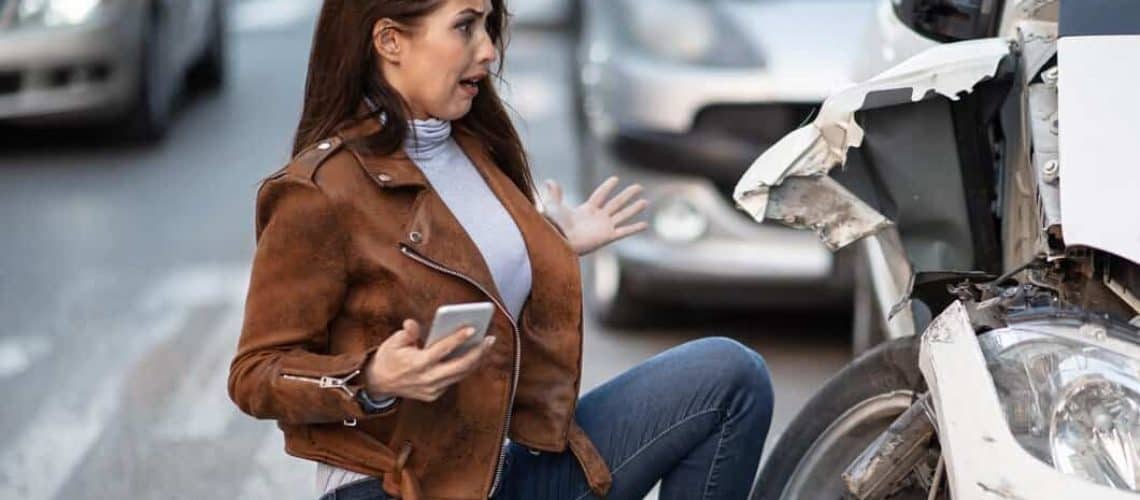 A woman holding a phone appears shocked, standing near a white car with front-end damage on a busy street.