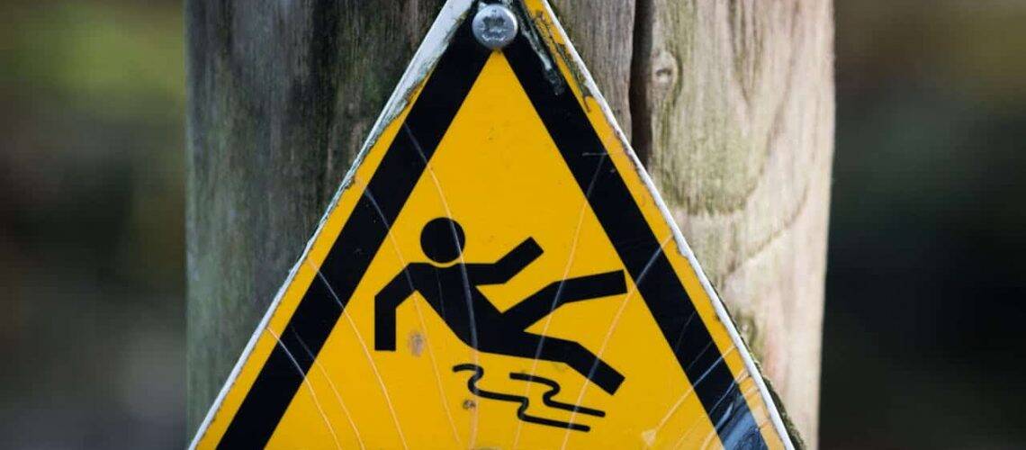 Yellow triangular sign on a wooden post warns of slippery surface, featuring a stick figure slipping.