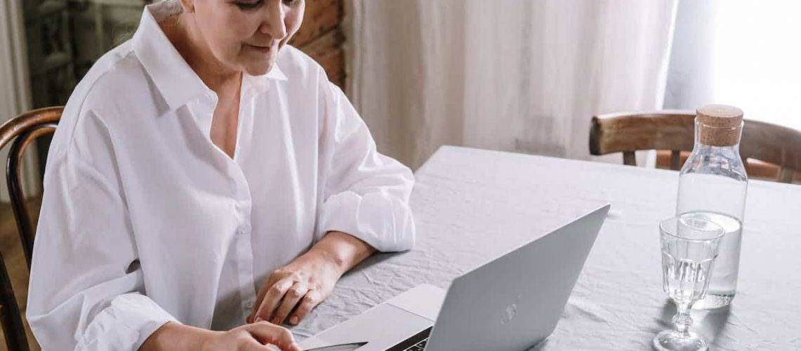 A person in a white shirt sits at a table using a laptop. A bottle and a glass of water are on the table.