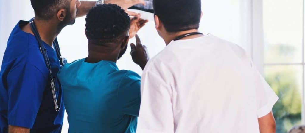 Three healthcare professionals in scrubs examine an X-ray together near a window.