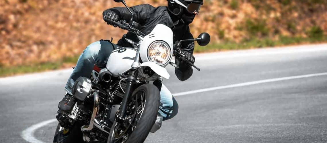 A person in a helmet and leather jacket rides a motorcycle on a curved road, leaning into the turn.