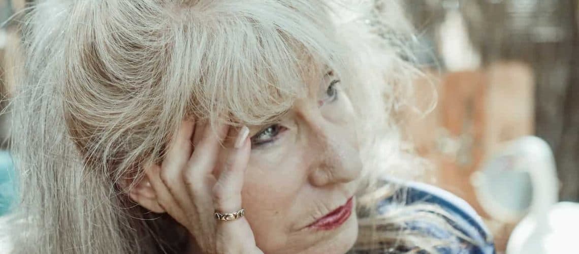 Older woman with long gray hair and a gold ring, resting her head on her hand, looking to the side. Background is blurred.