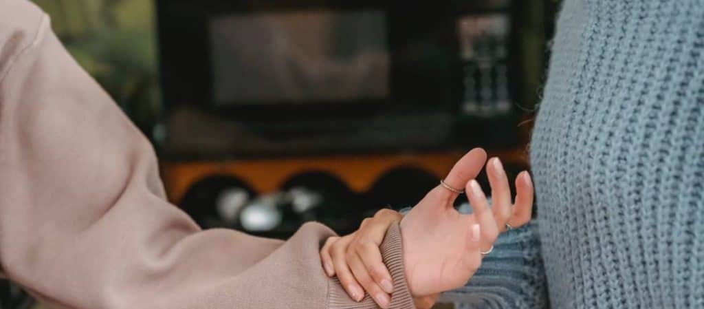 Two people stand close, one wearing a beige sweater gently holds the other's left wrist, who wears a blue textured sweater. Background shows a microwave and kitchen items.