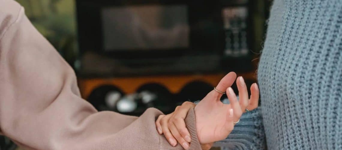Two people stand close, one wearing a beige sweater gently holds the other's left wrist, who wears a blue textured sweater. Background shows a microwave and kitchen items.