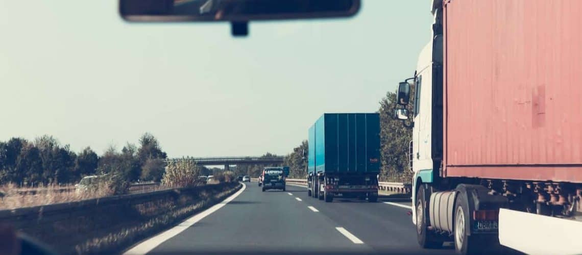 View from a car interior driving on a highway with three trucks ahead; blue sky and trees line the road.