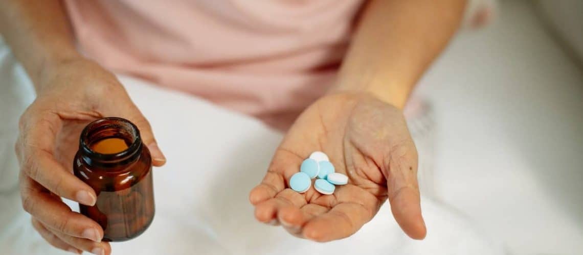 A person holds a brown pill bottle in one hand and a few blue and white pills in the other hand.