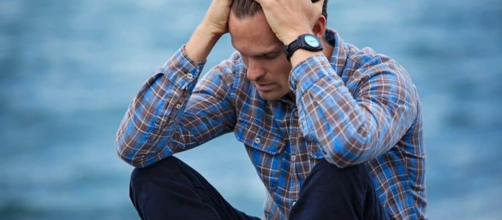 A person in a plaid shirt and watch sits by the water, holding their head with both hands, appearing pensive.
