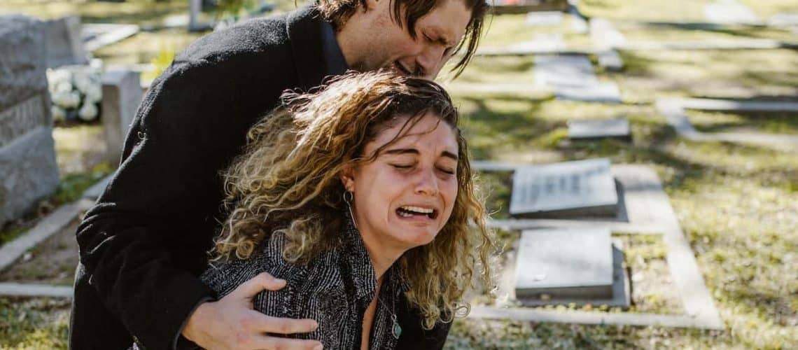 A woman is crying at a cemetery, supported by a man who appears to be comforting her. Headstones are visible in the background.