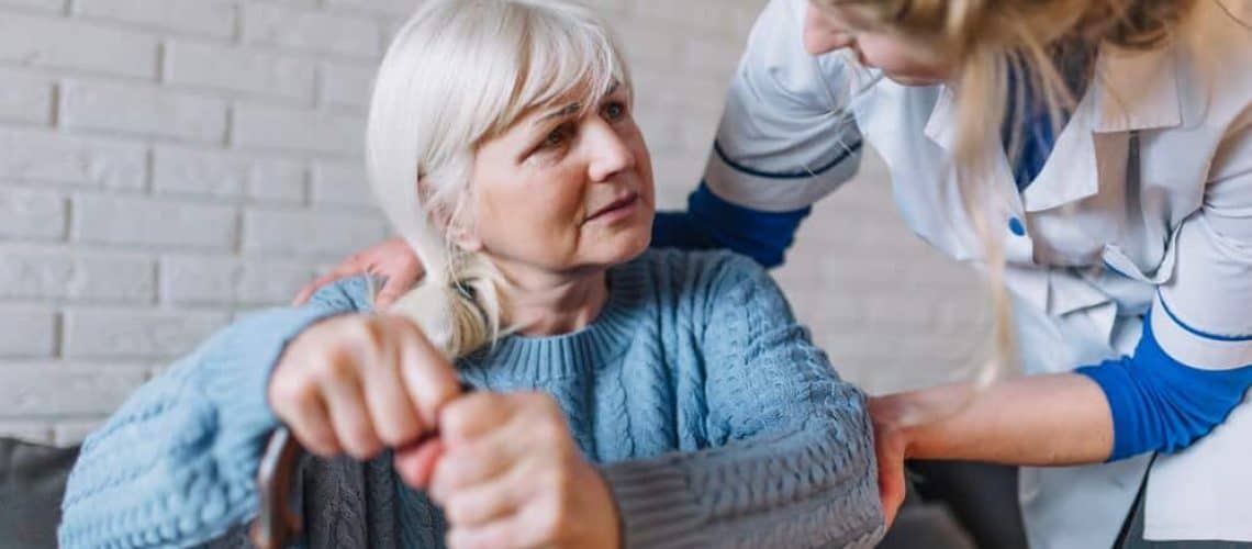 Elderly woman with white hair sitting and holding a cane, assisted by a nurse in a white uniform.