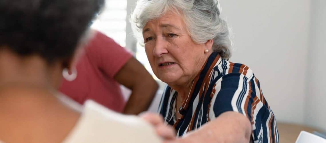 An elderly woman with gray hair, wearing a striped shirt, appears to be consoling a person in the foreground. Another person is partially visible in the background.